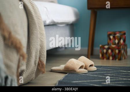 Komfortabel aussehendes Schlafzimmer mit weichen Hausschuhen, die auf Teppich neben Bett gelegt werden, und einem hölzernen Nachttisch mit farbenfrohen Kästen, die im Hintergrund sichtbar sind Stockfoto