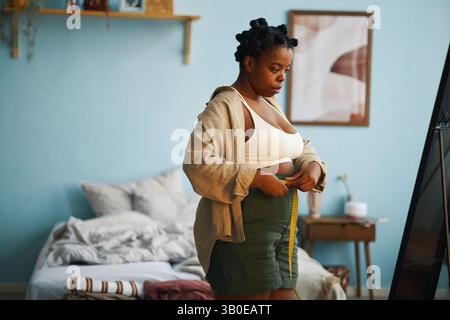 Afroamerikanische Frau, die sich in einem gemütlichen Schlafzimmer entspannt, neben dem Bett steht und ein Maßband hält, mit einem nachdenklichen Ausdruck Stockfoto