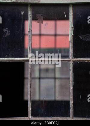 Altes Holzfenster auf einem alten verlassenen Gebäude mit reflektierendem Fenster und einem Gebäude mit einem roten Dach, das rechteckige Formen durch Fenster geschaffen hat Stockfoto