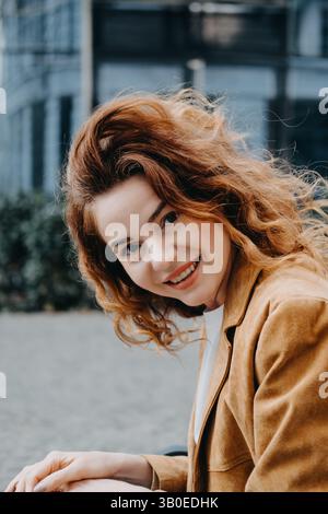 Eine junge Frau mit lockigen roten Haaren lächelt fröhlich, während der Wind ihr Haar vor das Gesicht wirft. Fröhliches Leben, spontanes Lächeln, Wohlfühlen Stockfoto