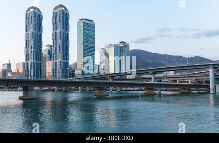 Busan, Südkorea - 16. März 2018: Modernes Stadtbild mit Wolkenkratzern, einer Brücke über ruhiges Wasser und einer bergigen Kulisse während der Dämmerung, Highl Stockfoto