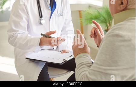 Seniorkranker Mann, der mit einem männlichen Arzt in weißer Uniform spricht und während der ärztlichen Untersuchung in der Klinik sitzt. Stockfoto
