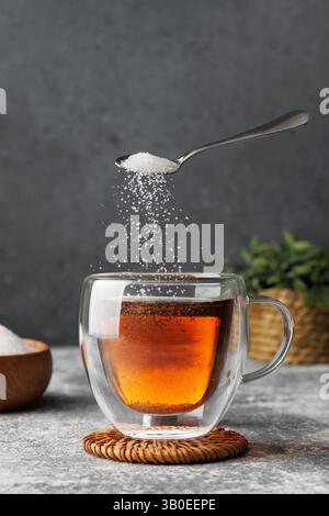 Weißer Zucker, der vom Teelöffel in eine Glasschale mit heißem Tee fällt, auf den Tisch vor grauem Hintergrund gestellt Stockfoto