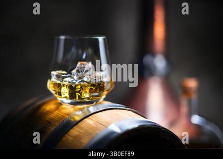 Ein Glas Whiskey mit Eiswürfeln auf altem Eichenfass. Verschwommene Flasche und Kupferalembic auf Hintergrund. Traditionelles Alkoholdestilleriekonzept Stockfoto