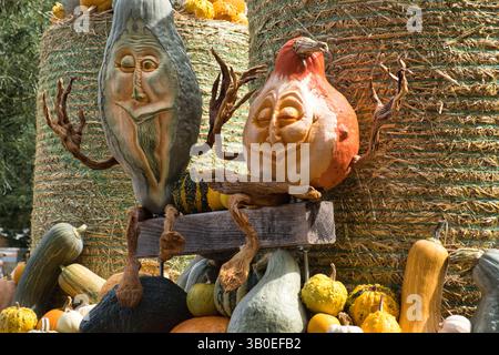 Zwei anthropomorphe Gemüsefiguren mit geschnitzten Gesichtern und zweigähnlichen Armen sitzen auf einer Holzkiste, umgeben von verschiedenen Kürbissen und Kürbissen. Stockfoto