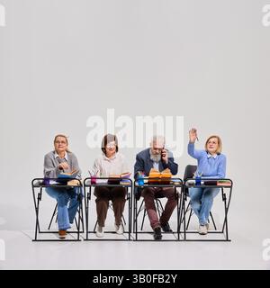 Gruppe von vier älteren Menschen als Schüler, die während des Unterrichts am Schreibtisch sitzen, Mann am Telefon sprechen, ältere Frau, die Hand zur Antwort hebt. Stockfoto