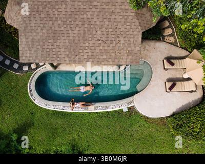 Zwei Personen schwimmen friedlich in einem ruhigen Pool, umgeben von üppigem Grün. Die Sonne scheint hell am klaren blauen Himmel und schafft ein perfektes Ambiente für Entspannung und Freizeit. Stockfoto