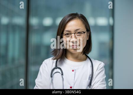 Porträt einer jungen asiatischen Ärztin. Der Doktor lächelt und blickt aus nächster Nähe in die Kamera. Die Krankenschwester in einem weißen medizinischen Mantel arbeitet im Büro einer modernen Klinik. Stockfoto