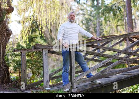 Bärtiger Mann in lässiger Kleidung posiert draußen auf einer verwitterten Brücke, umgeben von Bäumen und Sonnenlicht. Stockfoto