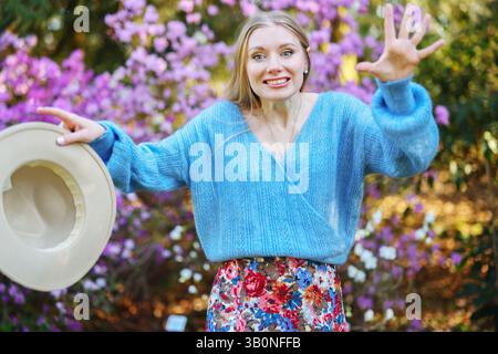 Überraschte Frau in blauem Pullover und Blumenrock reagiert in einem blühenden Garten spielerisch und hält einen Hut in der Hand. Stockfoto