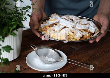 Karnevalssaison. Hausgemachte traditionelle italienische Karnevalsbonbons. Männliche Hände, die einen Teller mit Chiacchiere halten, mit Puderzucker bestreut, hölzerner Hintergrund. Stockfoto