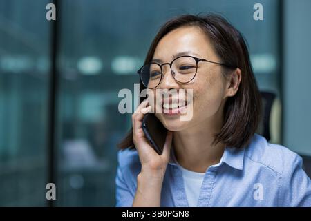 Junge erfolgreiche Geschäftsfrau, die im Büro in Nahaufnahme telefoniert. Zufriedene asiatische Frau lächelt, arbeitet am Fenster. Stockfoto