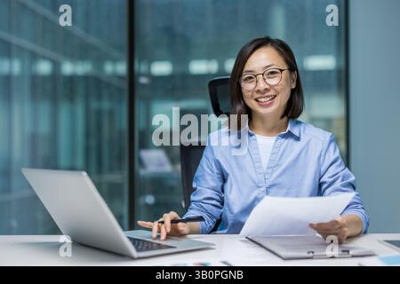 Porträt einer jungen erfolgreichen Geschäftsfrau Finanzierin am Arbeitsplatz im Büro. Asiatische Frau hinter Papierkram lächelnd und in die Kamera schauend, Frau, die Berichte ausfüllt und Steuerformulare vorbereitet. Stockfoto