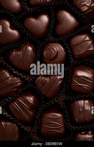 Eine reichhaltige Auswahl an handgefertigten dunklen Pralinen, elegant in schwarzen Pappbechern angeordnet, mit Herzformen, tropfenden Designs und seidenweichen Texturen. Stockfoto