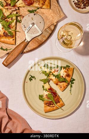 Gourmet-Pizza mit Rucola und Käse, serviert mit Weißwein auf einer Keramikplatte, neben einem hölzernen Schneidebrett und Pizzaschneider. Stockfoto