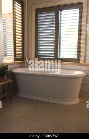 Freistehende weiße Badewanne im Badezimmer auf Steinboden unter Fensterläden, mit Topfpflanzen Stockfoto