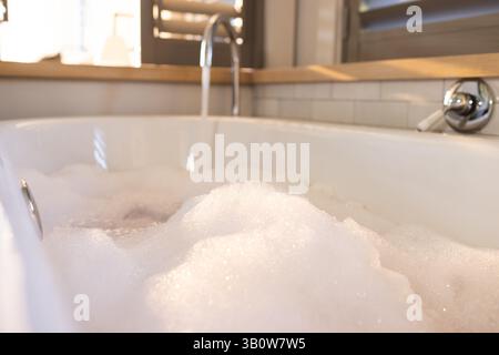 Die weiße Badewanne füllt sich mit Schaumblasen unter dem Chromhahn im Badezimmer, in der Nähe der Fensterläden Stockfoto