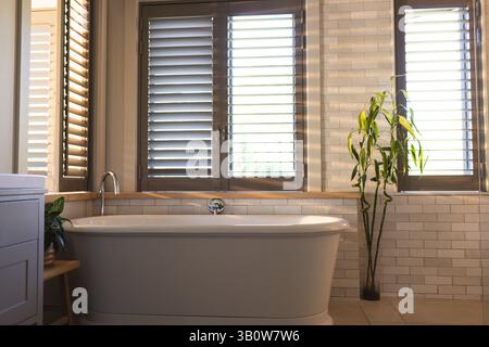 Weiße freistehende Badewanne im hellen Badezimmer mit Holzläden und grünen Pflanzen Stockfoto