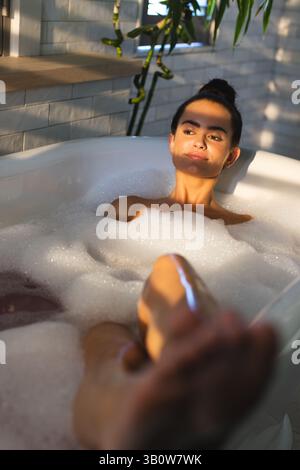 Entspannende Frau, die sich in einer freistehenden Badewanne im modernen Badezimmer mit Schaumblasen und Sonnenlicht zurücklehnen kann Stockfoto