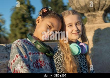 Zwei Mädchen, die Kopfhörer um den Hals tragen, posieren zusammen in einem Park, genießen die Sonne und genießen Musik, während sie lachen und las kreieren Stockfoto