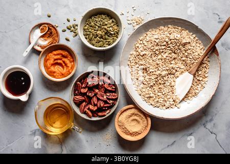 Eine Auswahl an Zutaten für ein Müsli-Rezept, einschließlich Hafer, Pekannüsse, Kürbispüree, Ahornsirup, Kürbiskerne und Gewürze, auf Marmor angeordnet Stockfoto