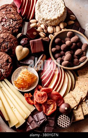 Eine Auswahl an Snacks mit roten Äpfeln, Keksen, Schokolade, getrockneten Früchten, Käsescheiben, und Nüsse auf einer Holzplatte. Stockfoto