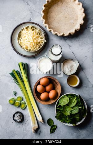 Eine Auswahl an Kochzutaten, darunter Lauch, Spinat, Eier und Milchprodukte, die auf einer Marmorarbeitsfläche angeordnet sind und bereit für die Rezeptvorbereitung sind Stockfoto