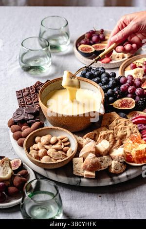 Eine Auswahl an Snacks, darunter Käsefondue, Brot, Cracker, Mandeln, Feigen, Trauben und Schokolade sind kunstvoll auf einem Tisch mit Glaswaren angeordnet. Stockfoto