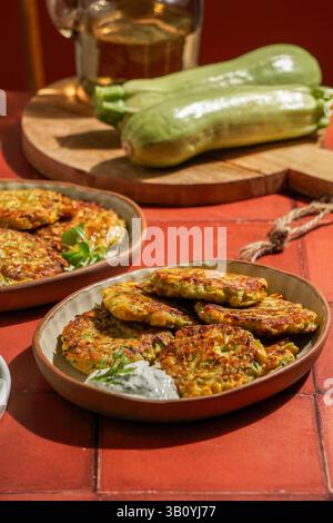 Zucchini-Krapfen auf Keramikplatten, begleitet von grünem Salat, cremiger Dippsauce, rustikaler Küche Stockfoto