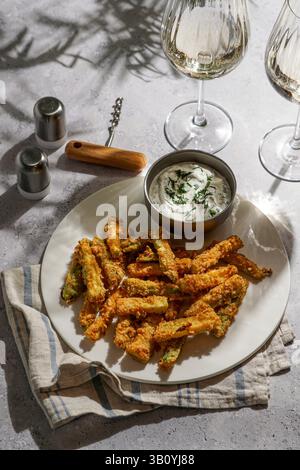 Gesunde knusprige Zucchini-Nuggets mit Sauce, auf grauem Steinhintergrund mit hellem Sonnenlicht, und ein Glas Weißwein Stockfoto
