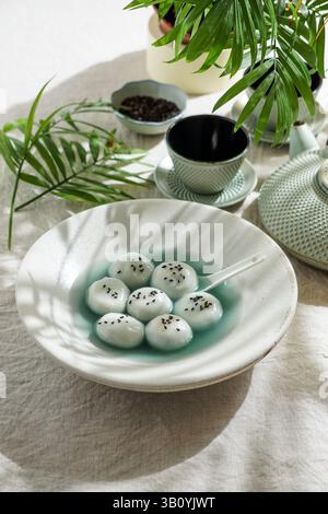 Tang Yuan, traditionelles asiatisches Dessert mit Glutinus-Reisbällchen in süßem Sirup, serviert in einer Schüssel mit einem Löffel, schwarzen Sesamsamen und Teekanne Stockfoto