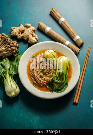 Schüssel mit Soba-Nudeln in scharfer Brühe mit Baby-Bok-Choy, umgeben von frischen Pilzen und Ingwer Stockfoto