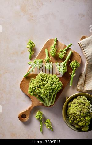 Ein Romanesco wird auf einem hölzernen Schneidebrett geschnitten, mit einem weiteren gewaschen und vorbereitet auf einem hellbraunen Hintergrund. Stockfoto