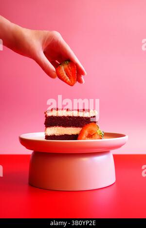 Weibliche Hand, die Erdbeere auf die Scheibe des zweilagigen roten Samtkuchens legt. Pop Art Food Color Blocking Ästhetik. Stockfoto