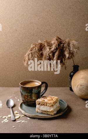 Hausgemachter norwegischer süßer Kuchen Verdens Beste Kake mit Vanillepudding, Meringue und Mandeln, ein quadratisches Stück auf Teller auf beigem Tisch mit Kaffeetasse, Löffel, Stockfoto