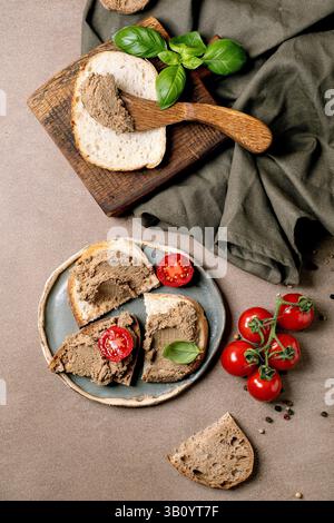 Hausgemachte Leberpastete auf Brotscheiben serviert mit frischen Kirschtomaten Basilikumblättern auf Holzbrett und Keramikplatte. Vorspeise im rustikalen Stil. Stockfoto