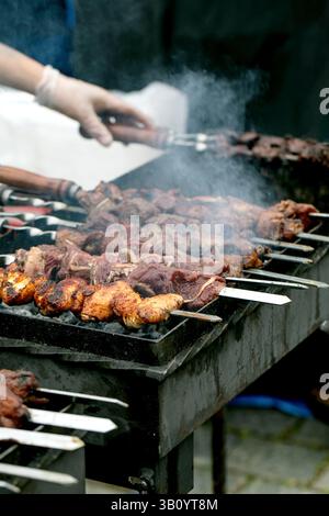 Person, die verschiedene Fleischspieße auf dem Grill im Freien grillt. Saftige Kebabs mit Hühnchen- und Rindfleischstücken, die auf Metallspießen gekocht werden und Rauch von h aufsteigt Stockfoto