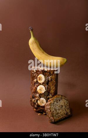 Modernes Stillleben mit hausgemachtem Bananenbrot. Brot mit Walnüssen, bestreichen Karamell, belegt mit frischer Banane auf braunem Hintergrund. Levitation Banane abo Stockfoto