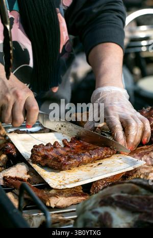 Street Food Festival in Europa. Traditionelle europäische gegrillte Schweinerippchen in Sauce. Der Verkäufer in Handschuhen bereitet eine Portion Rippchen am Essensstand vor. Stockfoto