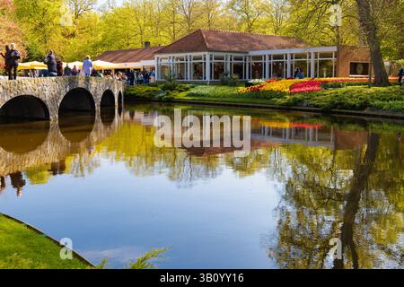 Keukenhof, Niederlande - 2023 - 05 - 02: Malerischer Blick auf die Gärten des Keukenhof mit einem Gebäude, einer Steinbrücke, Besuchern und farbenfrohen Tulpenbetten Stockfoto