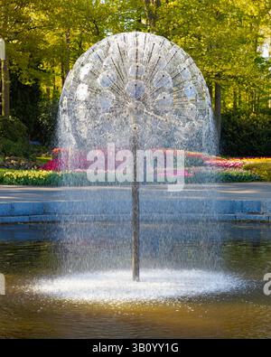 Moderner sphärischer Wasserbrunnen, der Tropfen in einem Teich in den Gärten des Keukenhof in den Niederlanden sprüht, mit bunten Blumenbeeten und Bäumen im Hintergrund. Stockfoto