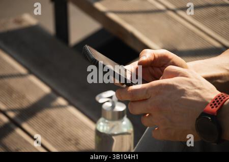 Smartphone in den Händen sportlicher Mann. Mann sitzt auf Treppen und überprüft Apps mit Wasserhaushalt nach dem Training bei Hitze. Sommersaison. Nahaufnahme. Stockfoto