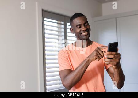 Lächelnder afroamerikanischer Mann, der in einem sanft beleuchteten Raum mit weißen Plantagenläden das Smartphone anzapft Stockfoto