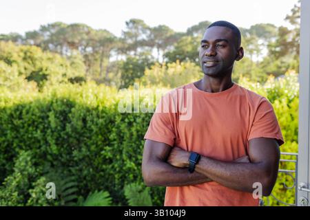 Stehender afroamerikanischer Mann, der seine Arme auf der Veranda zum Hinterhof kreuzt, mit Smartwatch, Kopierraum Stockfoto