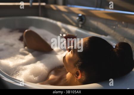 Liegende Frau, die sich in einer freistehenden Badewanne im Badezimmer mit Tee, Seifenblasen und Pflanzen entspannt Stockfoto