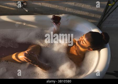 Liegende Frau, die sich in einer freistehenden Badewanne im Badezimmer mit Tee, Seifenblasen und Pflanzen entspannt Stockfoto