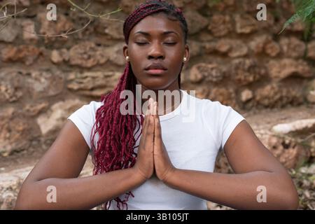Junge Frau mit Zöpfen, die Yoga praktiziert, meditiert mit ihren Händen zusammen und ihre Augen geschlossen vor einer Steinmauer in einem Park, was Well-bei fördert Stockfoto