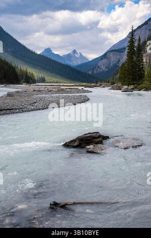 Ein ruhiger Fluss schlängelt sich durch ein üppiges Tal, umgeben von majestätischen Bergen unter einem dramatischen Himmel. Das klare Wasser spiegelt die lebendige Landschaft, cre, wider Stockfoto