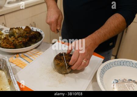 Eine Person schneidet gekonnt eine gebratene Artischocke gefüllt mit Kräutern in einer hellen Küche, während sie das Abendessen zubereitet. Stockfoto