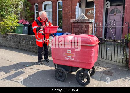 Ein Postbote der Royal Mail und sein Rollwagen für Pakete, die in einem Wohngebiet von Cardiff auf einer Lieferrunde verladen werden. April 2025. Stockfoto